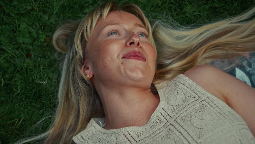 A carefree young blonde woman with blue eyes smiles and laughs with pure joy while lying in the lush green grass on a sunny summer day