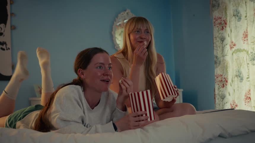 Two female friends lie on a bed eating popcorn while enjoying a movie night together in a cozy bedroom