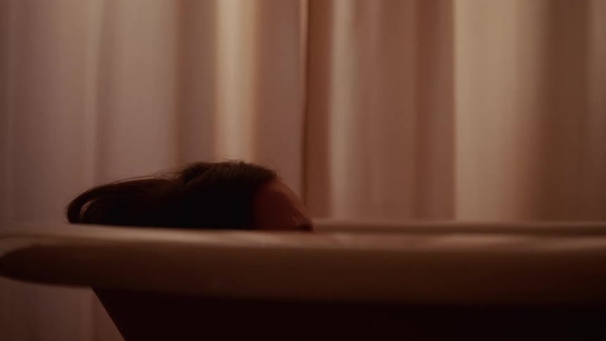 A somber woman with long dark hair slowly sinks below the water's surface in a bathtub