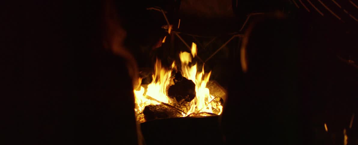 Two people are silhouetted against a roaring campfire at night, enjoying the warmth and dancing flames