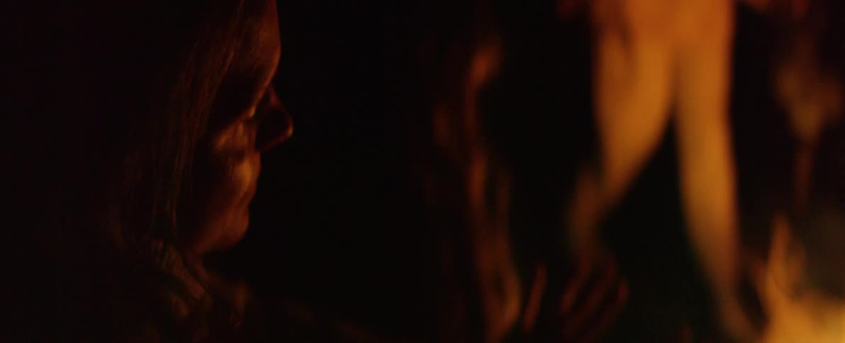 Side profile of an older woman's face illuminated by the warm, flickering glow of a fire at night