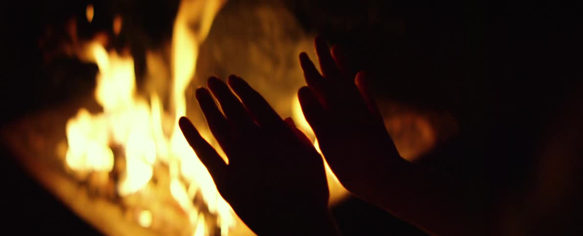 A person warms their silhouetted hands in front of a blazing campfire at night