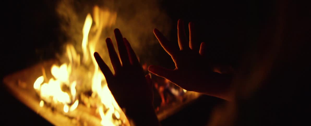 A person's hands are silhouetted as they warm them by a crackling bonfire at night