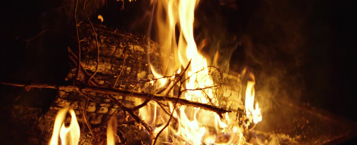 A close-up, slow-motion shot shows a campfire with bright orange flames consuming logs and twigs against a dark night background