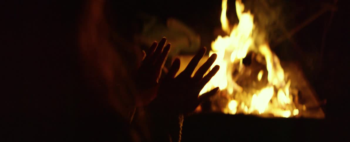 A person's silhouetted hands warming up in front of a crackling campfire at night in slow motion