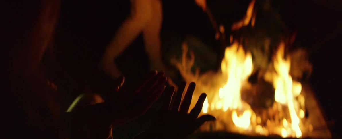 A person's face is illuminated by a cozy campfire before the shot reveals hands warming by the dancing flames at night