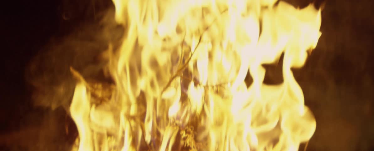 A close-up slow-motion shot shows bright yellow flames consuming kindling against a dark background