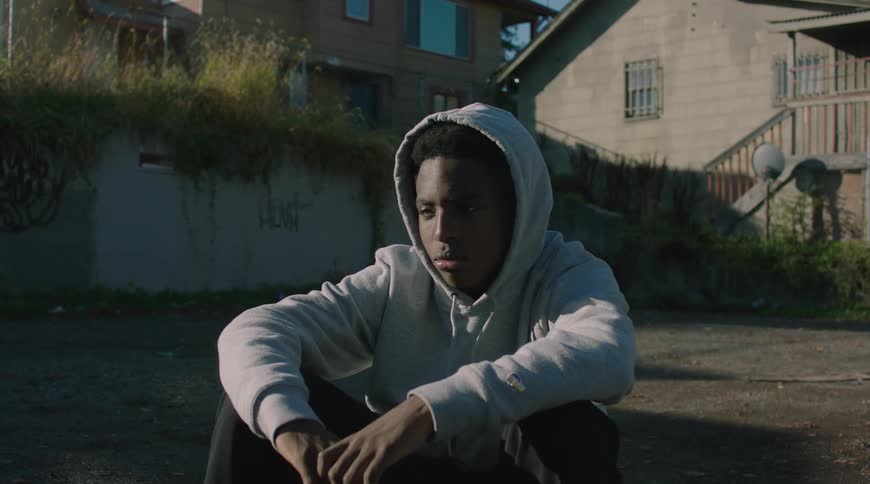 A thoughtful young Black man wearing a grey hoodie sits alone on the pavement in a quiet urban neighborhood, lost in thought