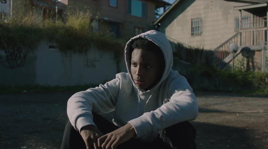 A somber young man in a grey hoodie sits alone in a rundown urban neighborhood, looking pensive