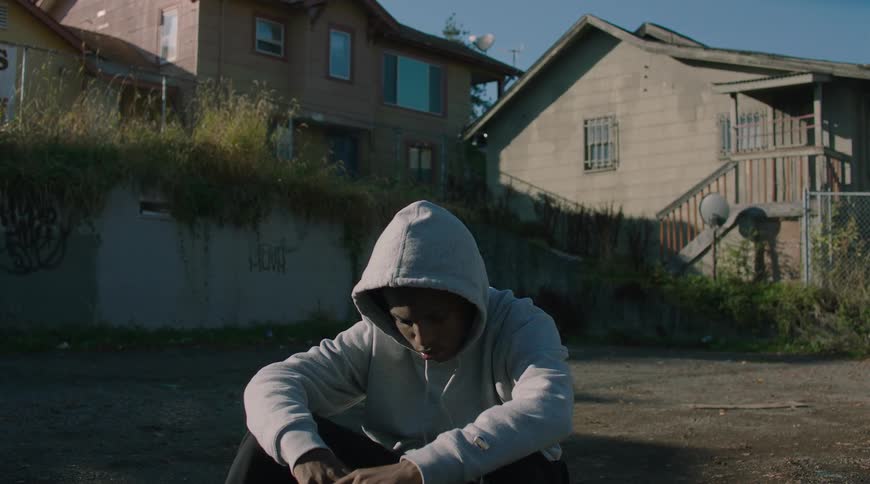 A slow zoom on a sad and contemplative young man in a grey hoodie sitting alone in a gritty urban neighborhood
