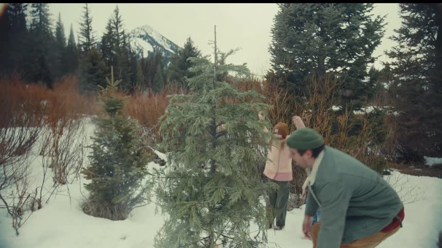 Excited children watch as their father hilariously fumbles and falls while cutting down their family Christmas tree in the snow