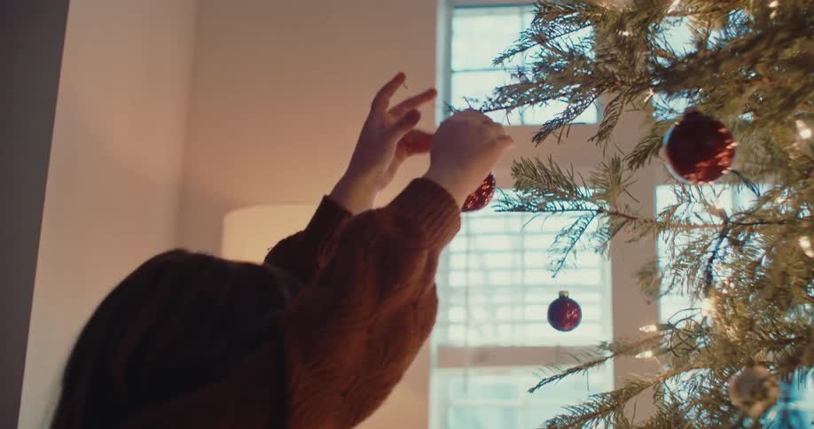 A happy little girl carefully hangs a red ornament on a lighted Christmas tree in her cozy home