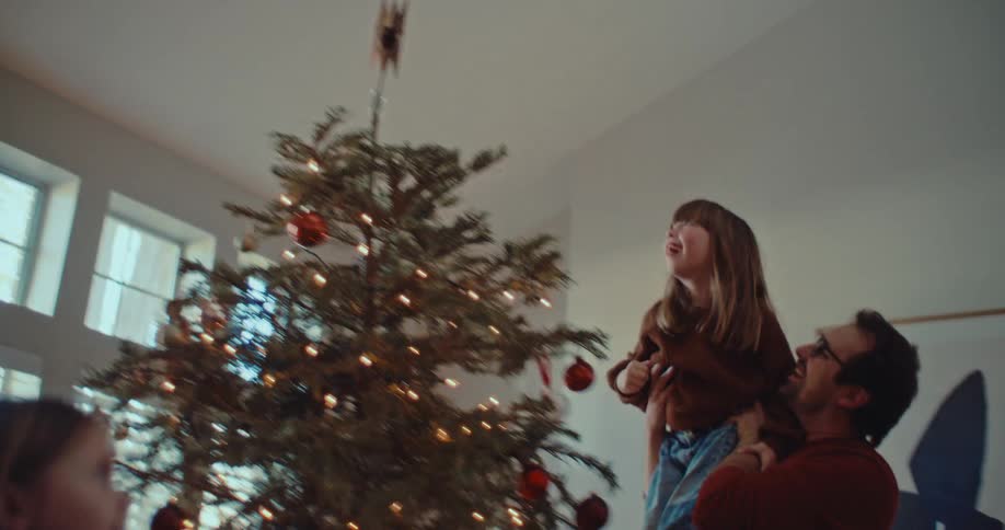 A happy father lifts his excited daughter to place the star on top of their Christmas tree while her sister watches in delight