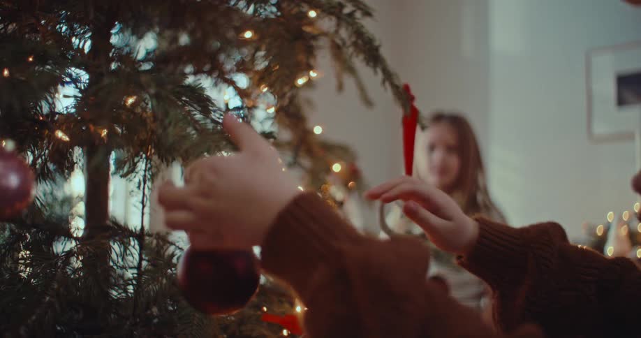 A young girl carefully hangs a purple ornament on a beautifully lit Christmas tree in slow motion, with her sister in the background