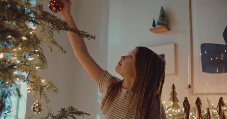 A young girl with wonder in her eyes carefully hangs a red ornament on the Christmas tree in a warmly lit home