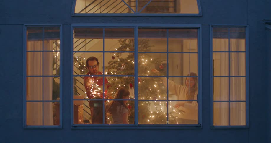 A cozy evening view through a window of a happy family decorating their Christmas tree together