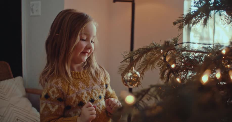 A joyful little girl with blonde hair carefully places a golden ornament on a warmly lit Christmas tree