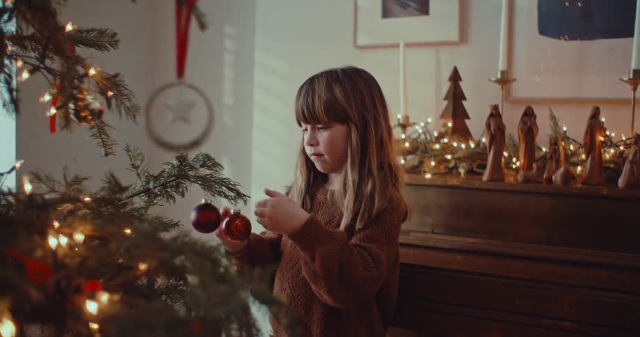 A focused little girl carefully hangs a red ornament on the Christmas tree and smiles with delight at her work