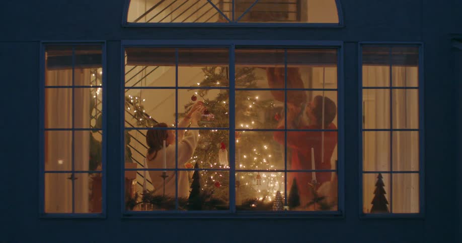 View from outside as a family joyfully decorates their glowing Christmas tree together inside their cozy home at night