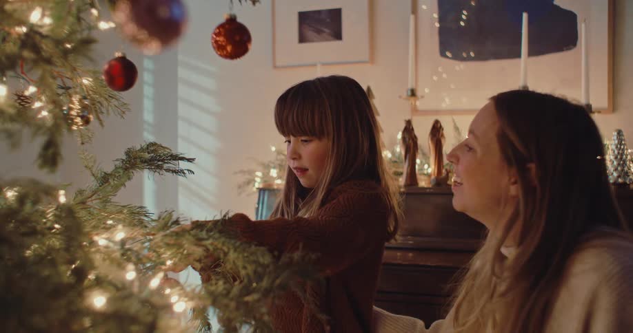 A loving mother and her young daughter share a tender moment decorating their Christmas tree together in a cozy, festively lit home