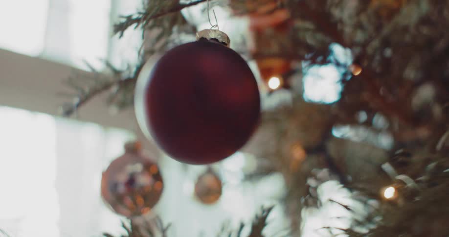 A close-up shot shows a red Christmas ornament being carefully removed from a box and hung on a festive, lit-up tree