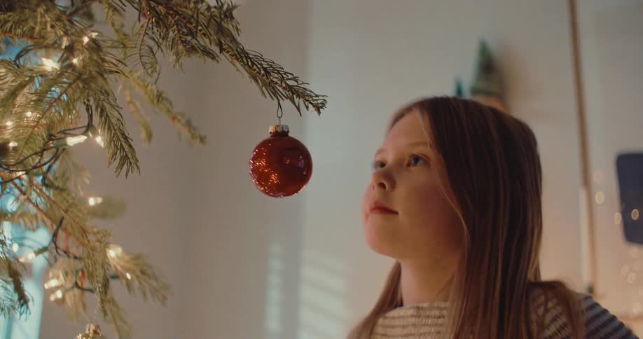 A young girl carefully places a shiny red ornament on a lighted Christmas tree and smiles as she admires it