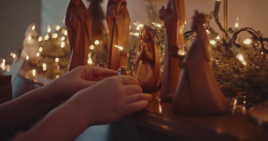 Child's hands place baby Jesus into a wooden nativity set surrounded by glowing Christmas lights