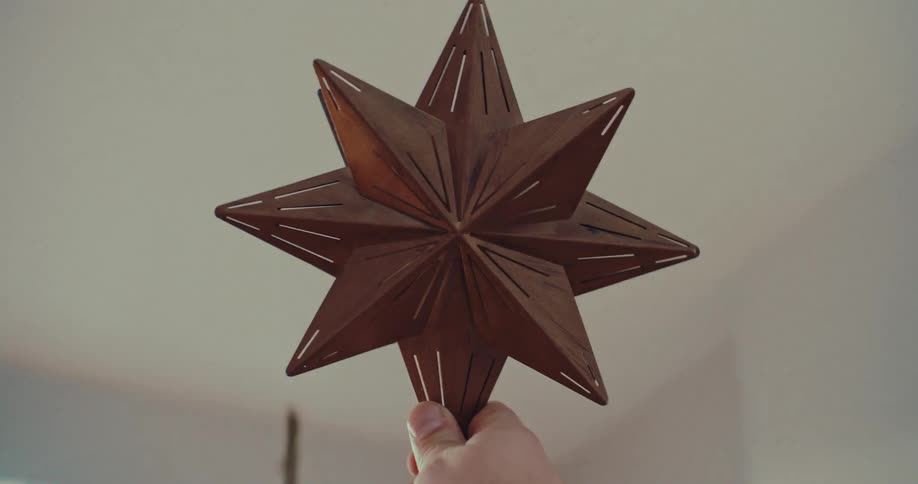 A hand lifts a star ornament from a box and places it atop a beautifully lit Christmas tree