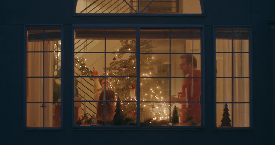 A joyous family with their daughter is seen through a brightly lit window decorating their Christmas tree for the holidays