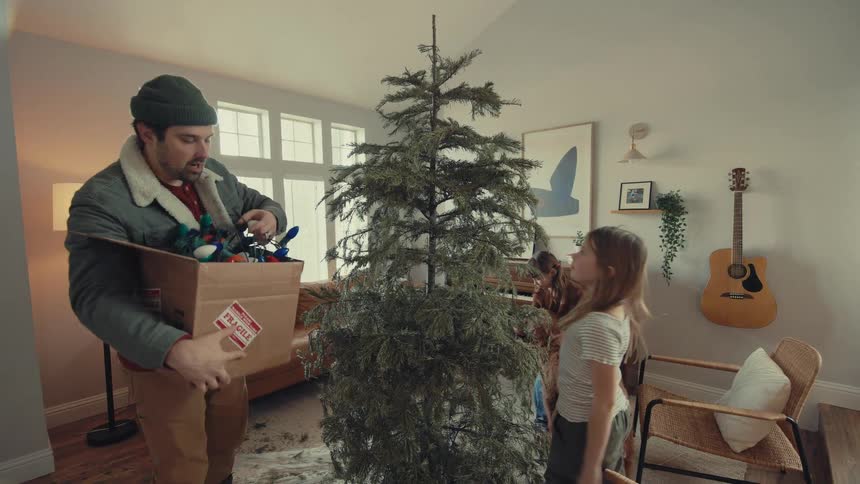 A happy family, including a father, two young daughters, and their dog, begin decorating a large Christmas tree together in their cozy living room