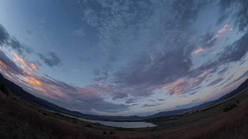 A serene fisheye timelapse captures the sunset sky with clouds passing over a peaceful mountain lake as day fades to night