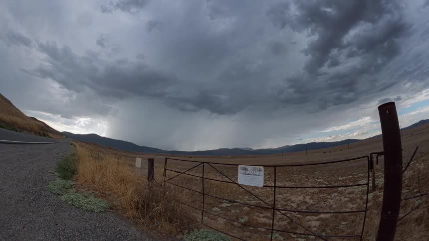 A dramatic wide-angle shot captures a storm system with heavy rain clouds moving over a vast, arid valley landscape