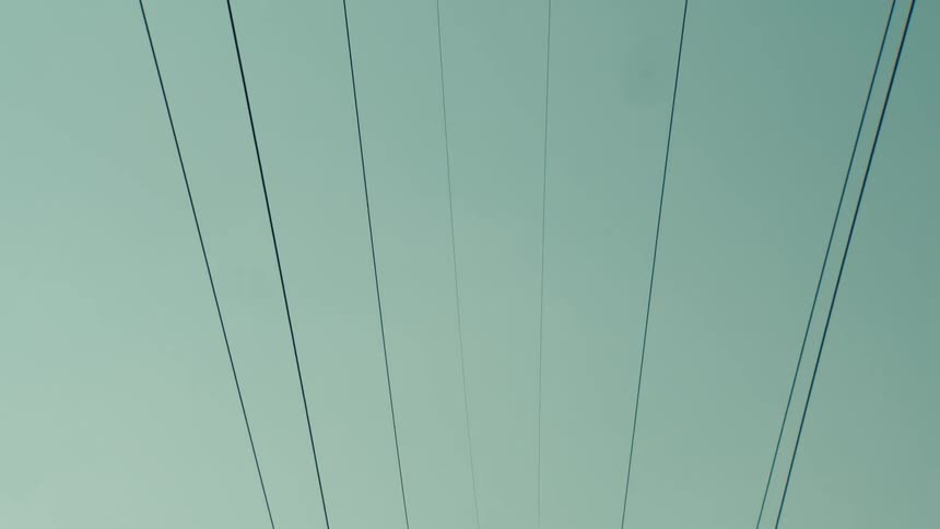 A slow tilt down from high voltage power lines reveals a large electricity pylon with a power plant cooling tower in the background