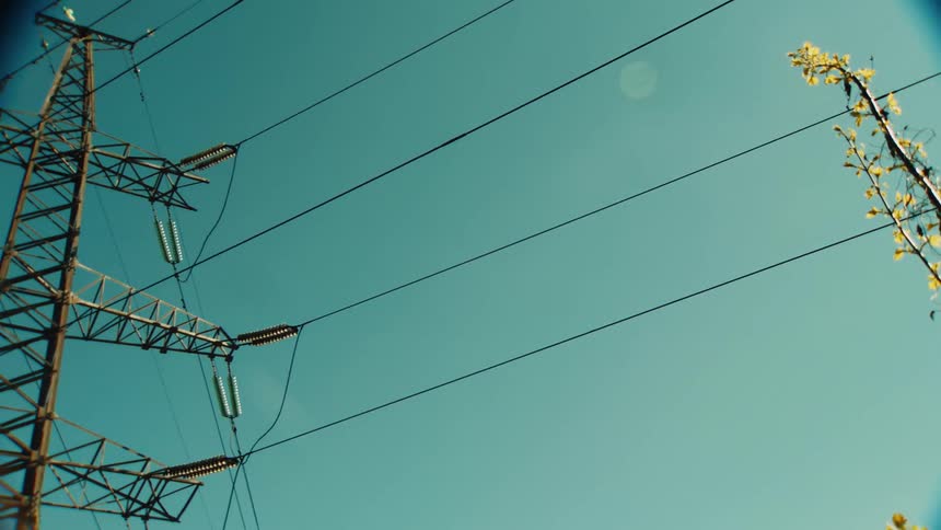 A vintage-style low-angle shot pans from budding tree branches to reveal a large electrical transmission tower against the clear blue sky