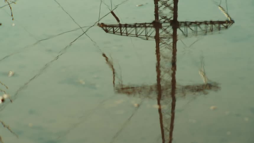 A high-voltage electricity pylon is reflected in the calm, rippling surface of the water