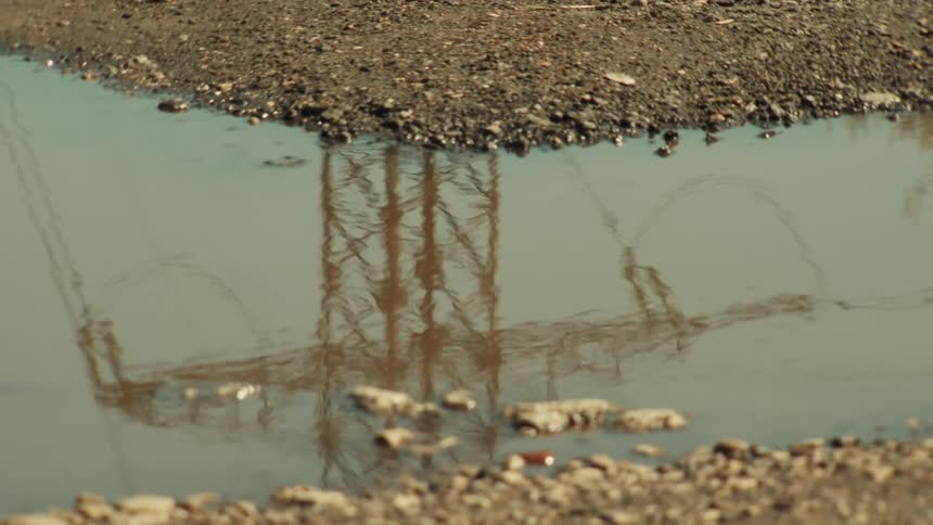 A slow zoom out reveals the reflection of an electricity pylon and power lines in a muddy puddle on a dirt road