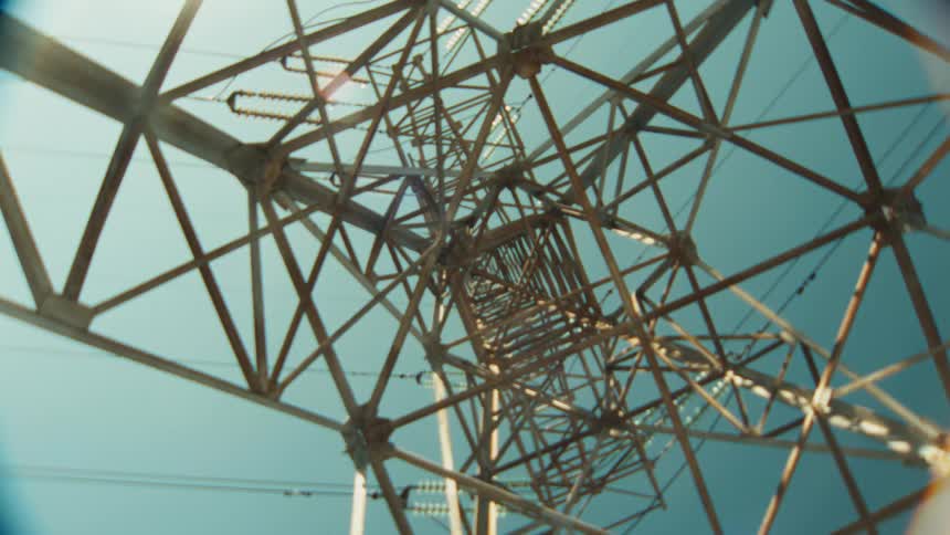 An abstract, low-angle shot slowly moving up through the geometric steel lattice of a transmission tower against a clear sky