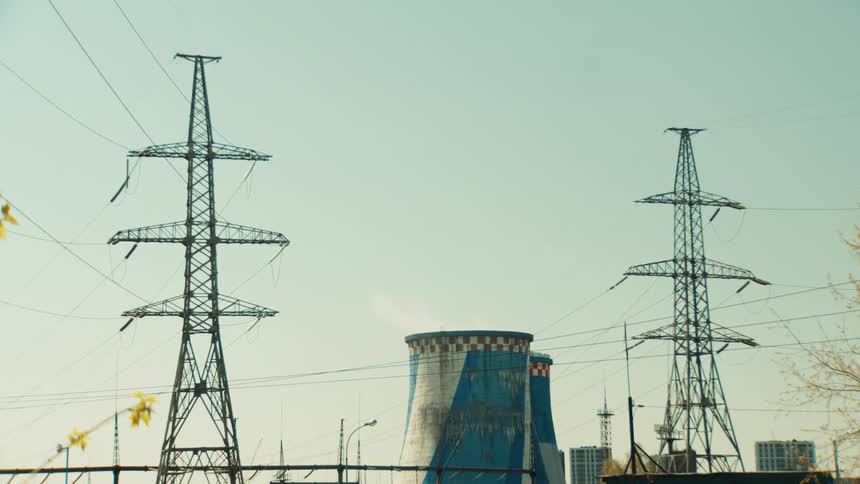 A power plant's cooling tower emits steam behind two large electricity pylons against a clear sky