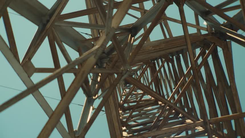 A low-angle zoom-out shot reveals the intricate, rusty metal structure of an electricity pylon against a clear blue sky
