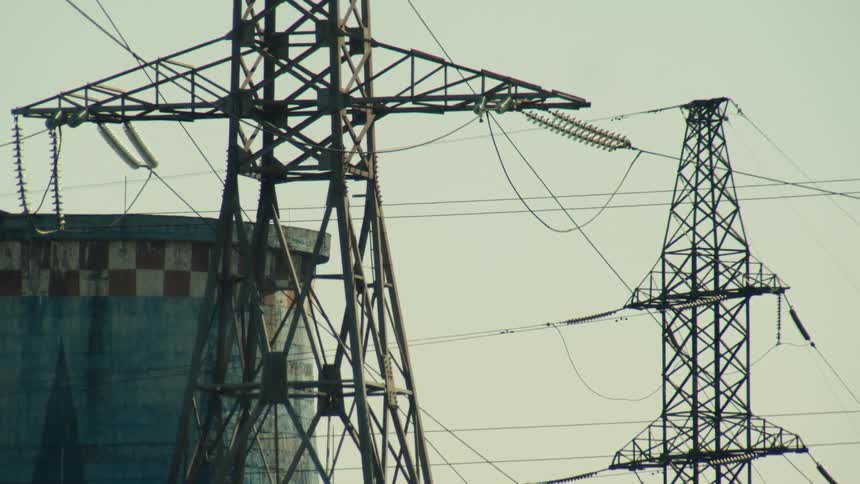 A static shot of massive high-voltage electrical towers and power lines next to an industrial plant