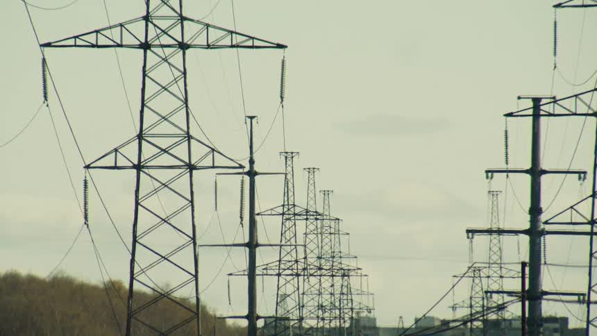 A static view of numerous high-voltage electricity transmission towers stretching into the distance under a pale, cloudy sky