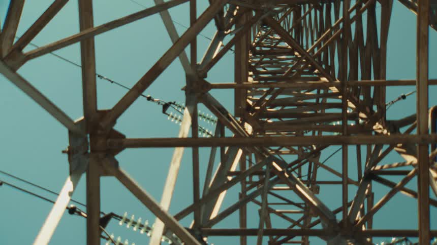 An upward-looking perspective reveals the intricate steel lattice of a high-voltage electricity pylon against a clear blue sky