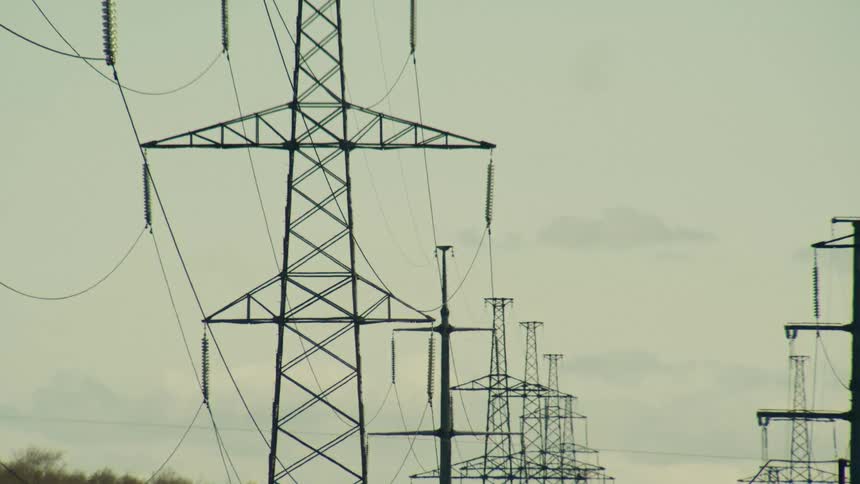 A row of high voltage power line towers stands against a hazy, overcast sky in a subtle time-lapse