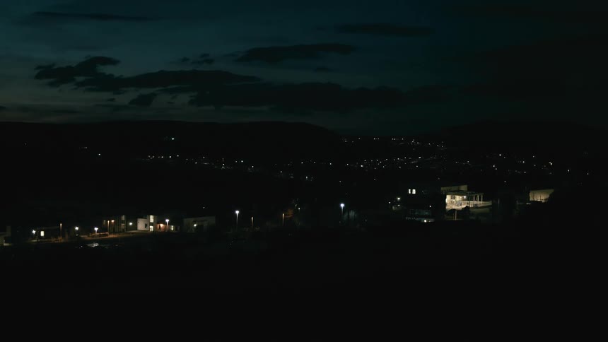 A still wide shot captures the quiet glow of a small town at night, with lights dotting the landscape against distant hills
