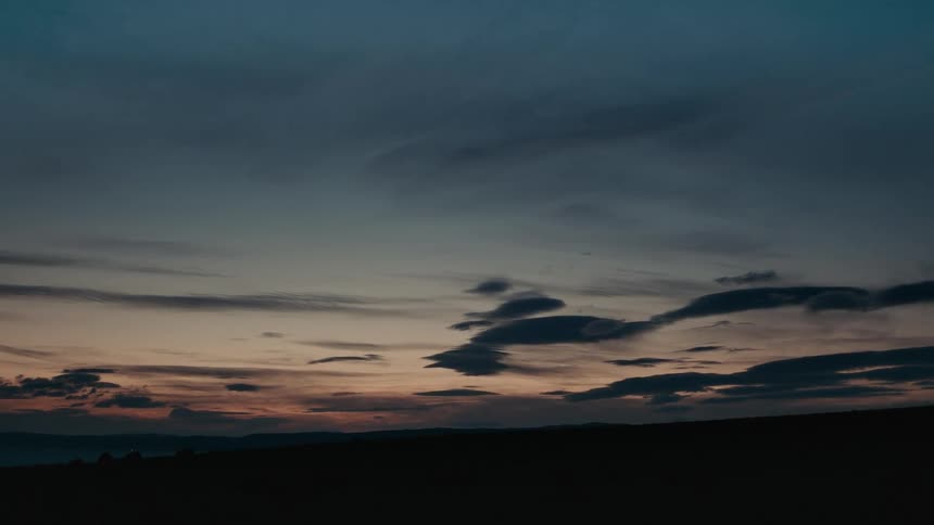 Dark clouds float peacefully across the twilight sky over a silhouetted landscape after the sun has set