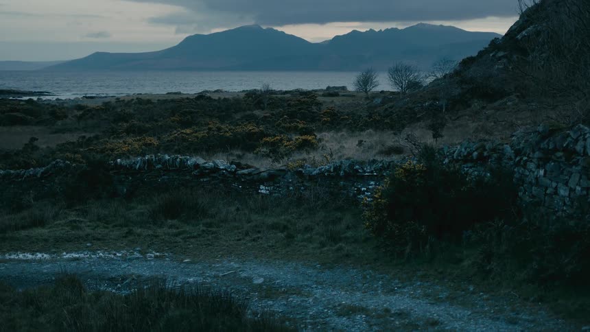 A desolate view of a rugged shoreline with a stone wall overlooking a vast loch and distant mountains on a cloudy day