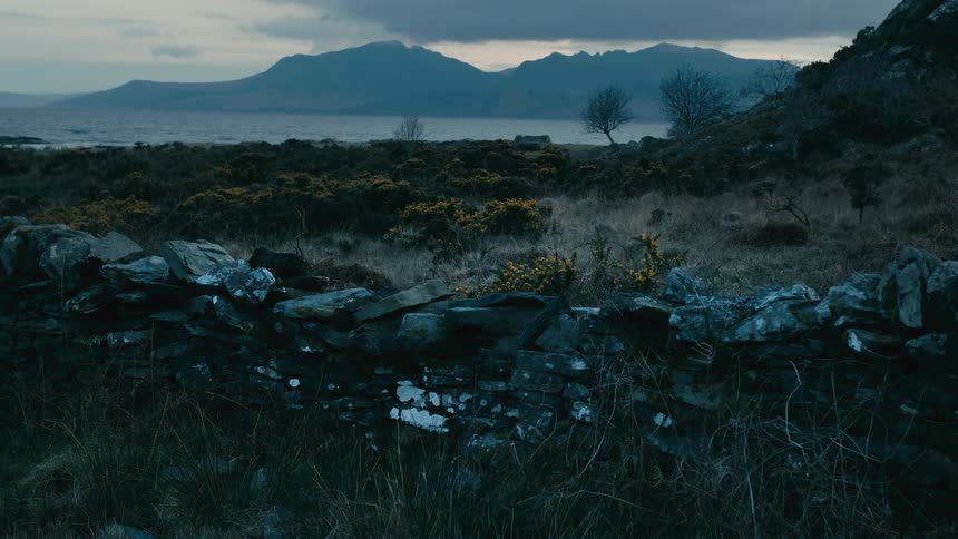 A rugged coastal landscape features a mossy stone wall in the foreground with a vast loch and mountains in the distance under a dramatic, cloudy sky