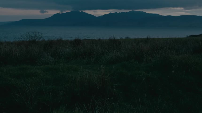 A still, cinematic view of a dark mountain silhouette across a calm sea at dusk