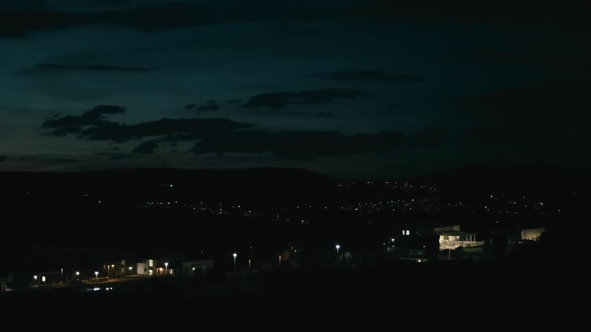A quiet town in a valley at night with streetlights and distant city lights under a dark, cloudy sky