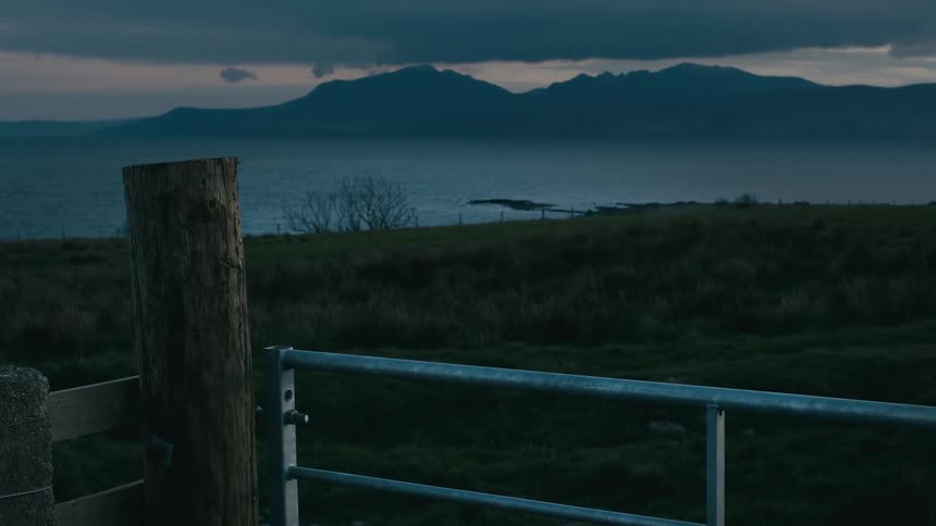 A serene and moody landscape view of a mountain range across a sea at dusk from a rural field